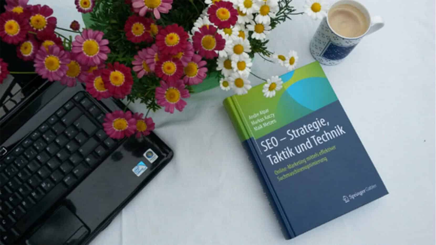 Komposition mit Notebook, Kaffeetasse, Blumenstrauß und SEO Strategie Buch